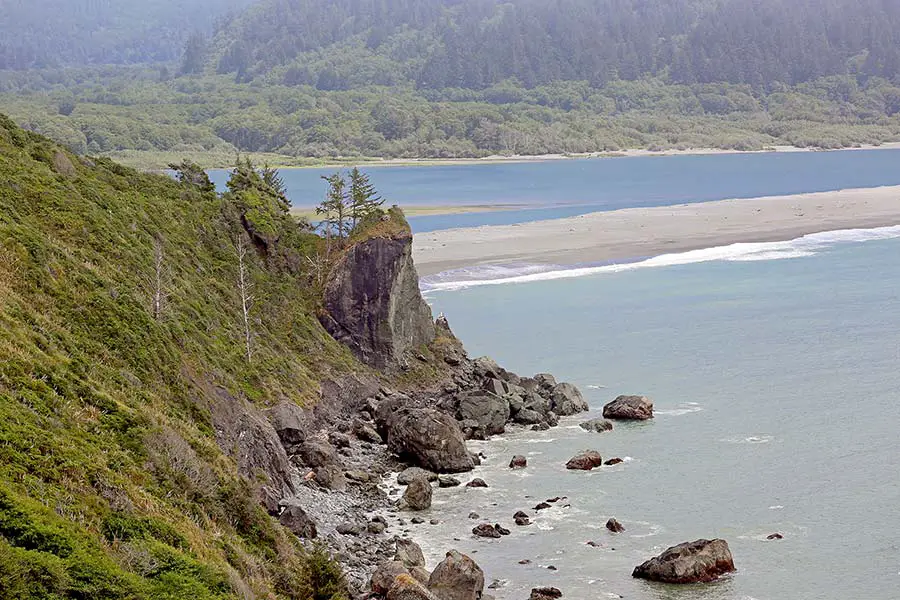 Steep mountain sidehill ends with huge boulders that line the Pacific Coastline, dense forest in background