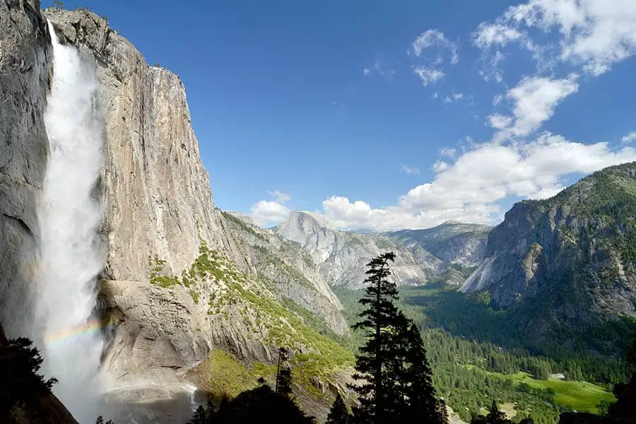 Upper water fall with rainbow in mist, distant view of forested Yosemite Valley surrounded by majestic mountains