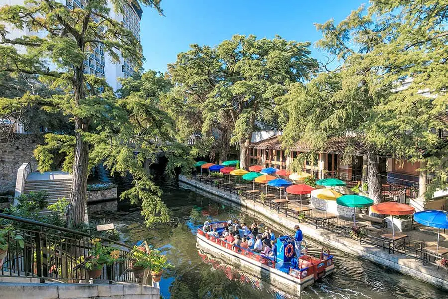 Boat carries sightseers past riverfront restaurant with outside dining under colorful umbrellas